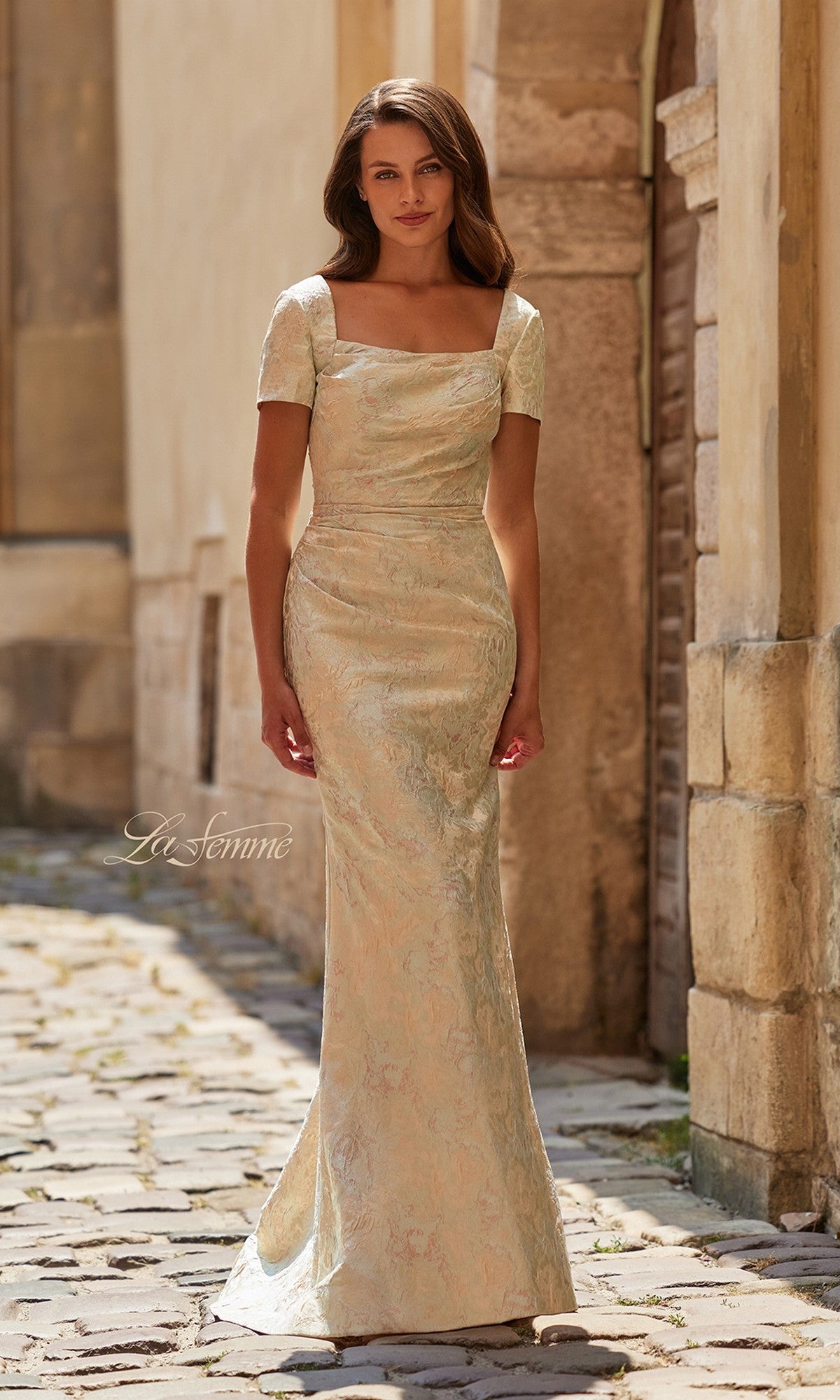 A woman stands on a cobblestone street wearing the La Femme 33598 long formal dress in a fitted, short-sleeve design with a square neckline and multi-color metallic floral brocade—perfect for any special evening occasion.
